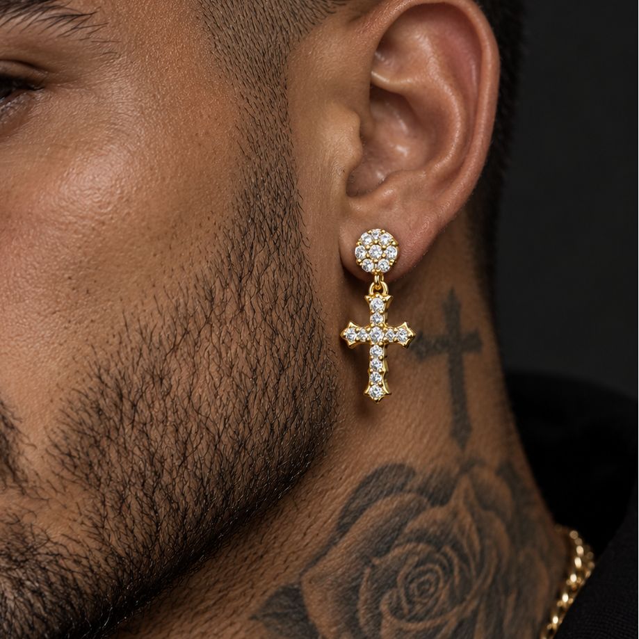 A close-up of a man's ear and neck wearing a gold cross drop earring adorned with clear crystals, along with a gold chain necklace and a black shirt.