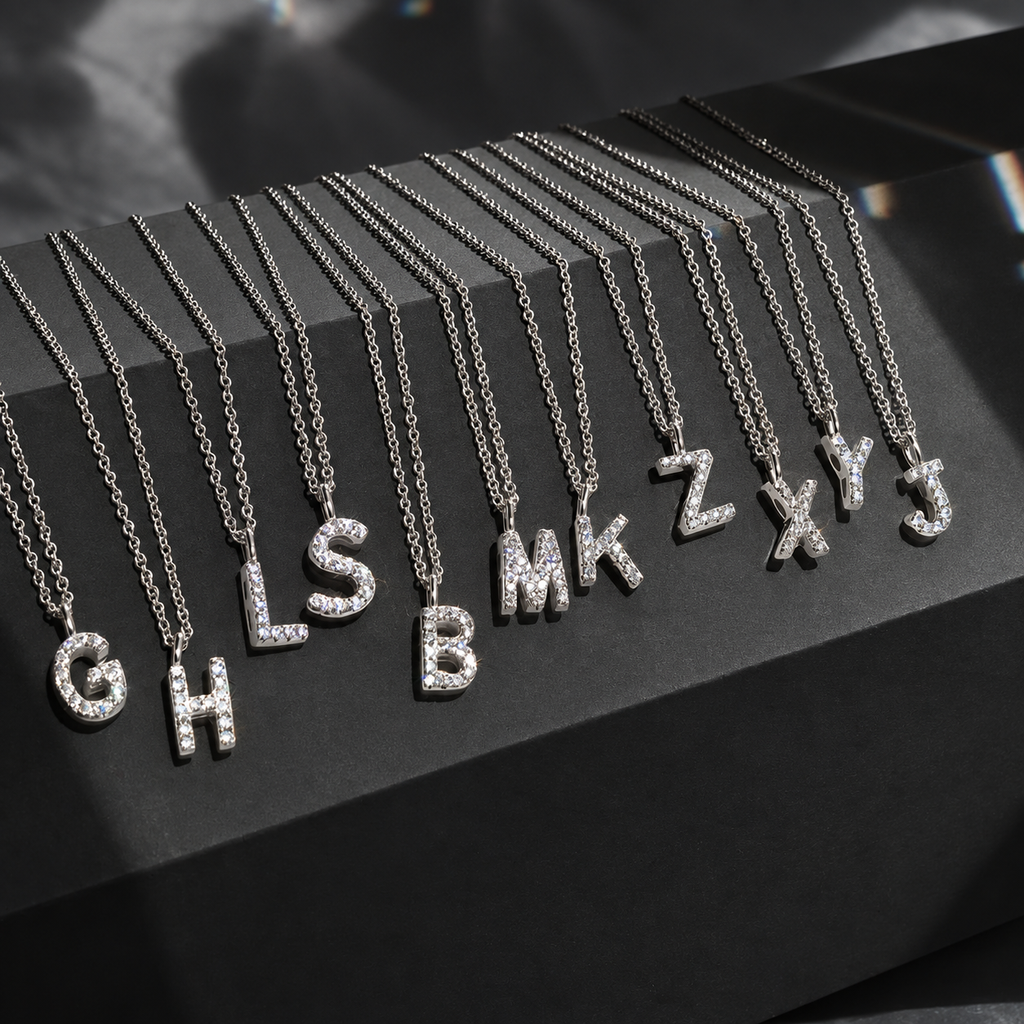 A set of silver letter necklaces with clear rhinestones, arranged in a fan shape on a black display block. The necklaces spell out the alphabet, starting with G, H, I, L, S, B, M, K, Z, X, Y, and J.