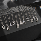 A set of silver letter necklaces with clear rhinestones, arranged in a fan shape on a black display block. The necklaces spell out the alphabet, starting with G, H, I, L, S, B, M, K, Z, X, Y, and J.